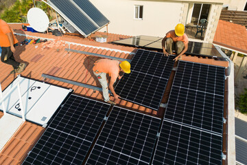 Workers install solar photovoltaic panels on the tiled terracotta roof of a house. Green energy...