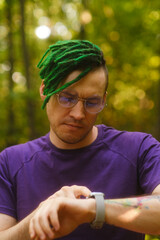 Fototapeta premium Man using smart watch in park. Male in violet t shirt with glasses and green deadlocks checking notifications on smart watch on summer day in park