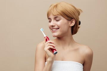an attractive, elegant woman stands on a beige background holding a red electric toothbrush in her hand and closes her eyes smiling