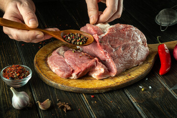 The cook sprinkles raw meat with dry pepper. Preparing beef meat before roasting. Working environment in the kitchen of a restaurant or cafe