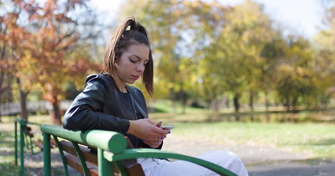 Beautiful girl sitting on a bench in the park. Fashionable young lady taking selfy photo with her phone.