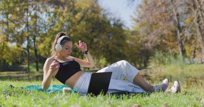 Beautiful girl with laptop and a coffee cup putting headphones on. Fashionable young lady lying in the park.
