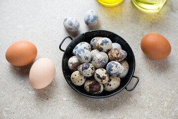 Black serving pan with raw quail and chicken eggs on a beige stone background, horizontal shot,...