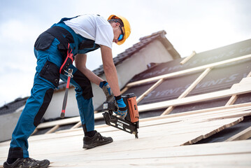 The roofer is nailing wooden battens on the roof with electric nailer. - obrazy, fototapety, plakaty