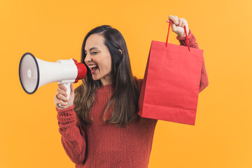 Latin-American cheerful girl holding a paper package bag after shopping and announcing sale in a megaphone, studio medium shot orange background. High quality photo