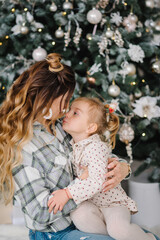 Baby girl, daughter hugs mom on the sofa near Christmas tree in the home. Happy New Year and Merry Christmas. Christmas decorated the interior of the house. The concept of a family holiday.