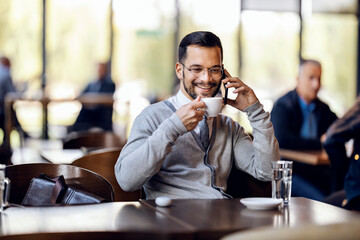 A happy young man is talking on the phone in coffee shop while sipping coffee.