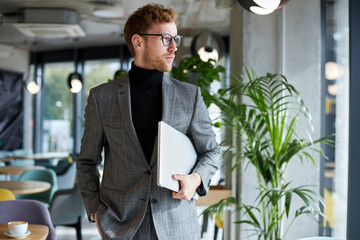 Young handsome businessman wearing stylish suit and eyeglasses holding laptop looking away, copy...