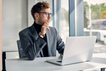 Portrait of young pensive businessman using laptop computer planning startup looking away in modern office. Handsome stylish freelancer wearing eyeglasses working from home 