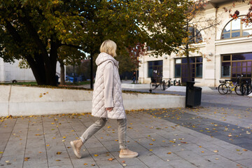 Alone blonde woman wearing white jacket walking at street. Side view. Concept of psychology and loneliss