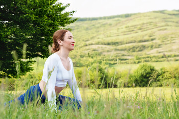 Close up portrait woman in sporty outfit exercising and stretching on mat in meadow outdoors. Hills landscape on background. Healthy active lifestyle