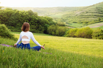 Naklejka na ściany i meble Young woman in sporty outfit relaxing and meditating on mat in sunset meadow outdoors. Hills landscape on background. Healthy active lifestyle in summer
