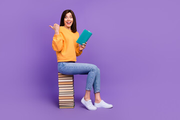 Full body length photo of smart intelligent girl sitting books pile excited hold literature finger pointing empty space crazy ad isolated on purple color background