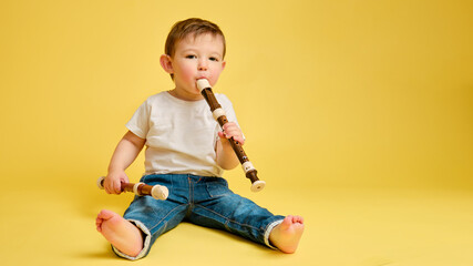 Toddler baby plays the flute, a child with a wind musical instrument on a studio yellow background. Happy child musician playing block flute, copy space. Kid aged one year and four months