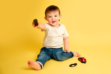 Toddler baby plays with toy cars on a studio yellow background. Happy child in white t-shirt and...