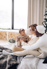 Happy interracial family sits on bed and plays with their baby daughter against background of Christmas tree and window.