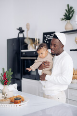 Happy african man holds his mixed race baby daughter on kitchen near Christmas decorations and pastry.