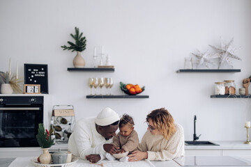 Happy interracial family plays with their baby daughter on kitchen near Christmas decorations.