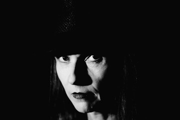 Black and white close up portrait against black background of woman 50plus with hat and looking...