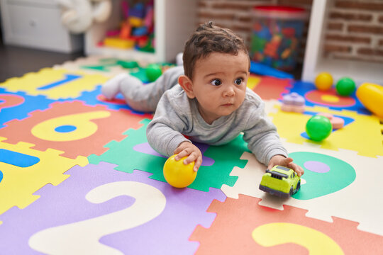 Adorable hispanic baby playing with ball and car lying on floor at kindergarten