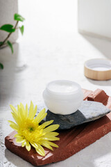 Obraz na płótnie Canvas White cream jar on natural stones podium and yellow flower on white background with light and shadows. Organic spa cosmetic beauty product