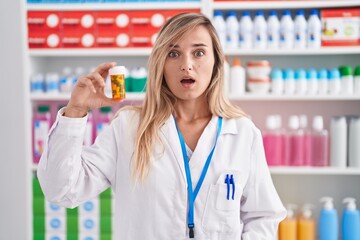 Young blonde woman working at pharmacy drugstore holding pills scared and amazed with open mouth...