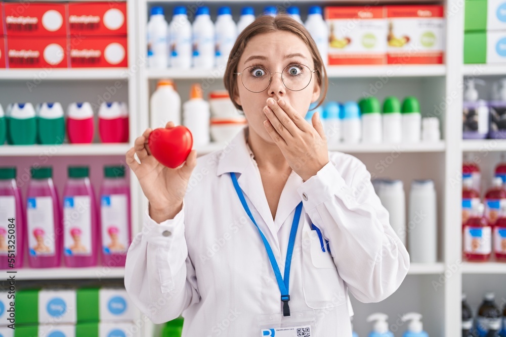 Sticker young caucasian woman working at pharmacy drugstore holding red heart covering mouth with hand, shoc