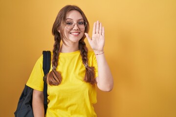 Young caucasian woman wearing student backpack over yellow background waiving saying hello happy and smiling, friendly welcome gesture