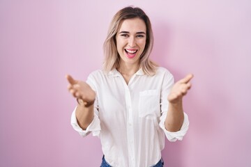 Young beautiful woman standing over pink background smiling cheerful offering hands giving assistance and acceptance.