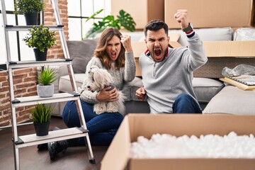 Young hispanic couple sitting on the floor at new home with log annoyed and frustrated shouting with anger, yelling crazy with anger and hand raised