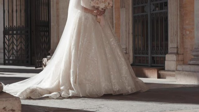 Gorgeous bride in fashion lace dress with long satin veil standing against ancient architecture background, fiancee holding charming wedding bouquet in hands waiting for her beloved groom. Elegant and