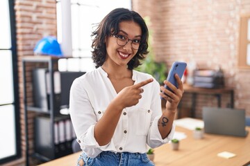 Young hispanic woman working at the office using smartphone smiling happy pointing with hand and finger