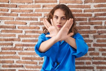 Beautiful brunette woman standing over bricks wall rejection expression crossing arms and palms doing negative sign, angry face
