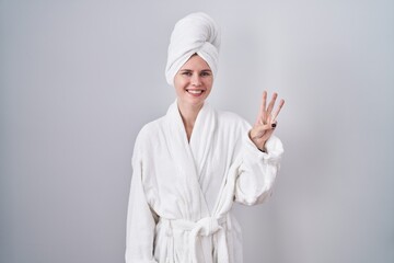 Blonde caucasian woman wearing bathrobe showing and pointing up with fingers number three while smiling confident and happy.