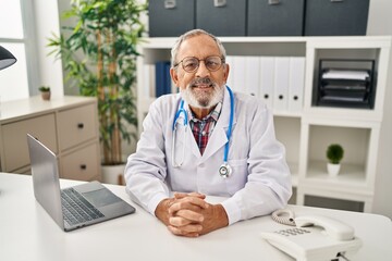 Senior grey-haired man doctor smiling confident sitting on table at clinic