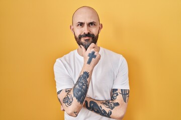 Young hispanic man with tattoos standing over yellow background looking confident at the camera...