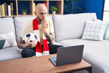 Hispanic man with tattoos watching football match hooligan holding ball on the laptop covering...