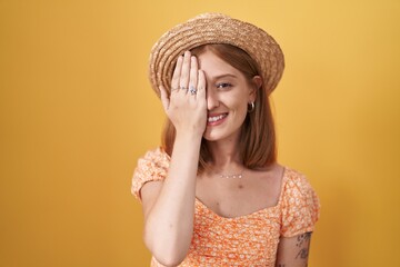 Young redhead woman standing over yellow background wearing summer hat covering one eye with hand, confident smile on face and surprise emotion.