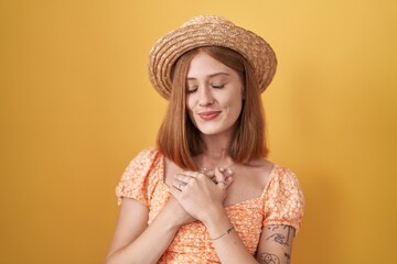Young redhead woman standing over yellow background wearing summer hat smiling with hands on chest with closed eyes and grateful gesture on face. health concept.
