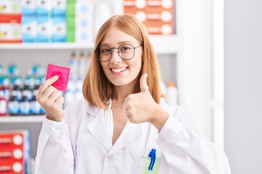 Young redhead woman working at pharmacy drugstore holding condom smiling happy and positive, thumb up doing excellent and approval sign
