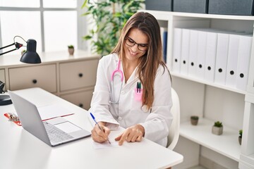 Young beautiful hispanic woman doctor using laptop writing on covid certificate card at clinic