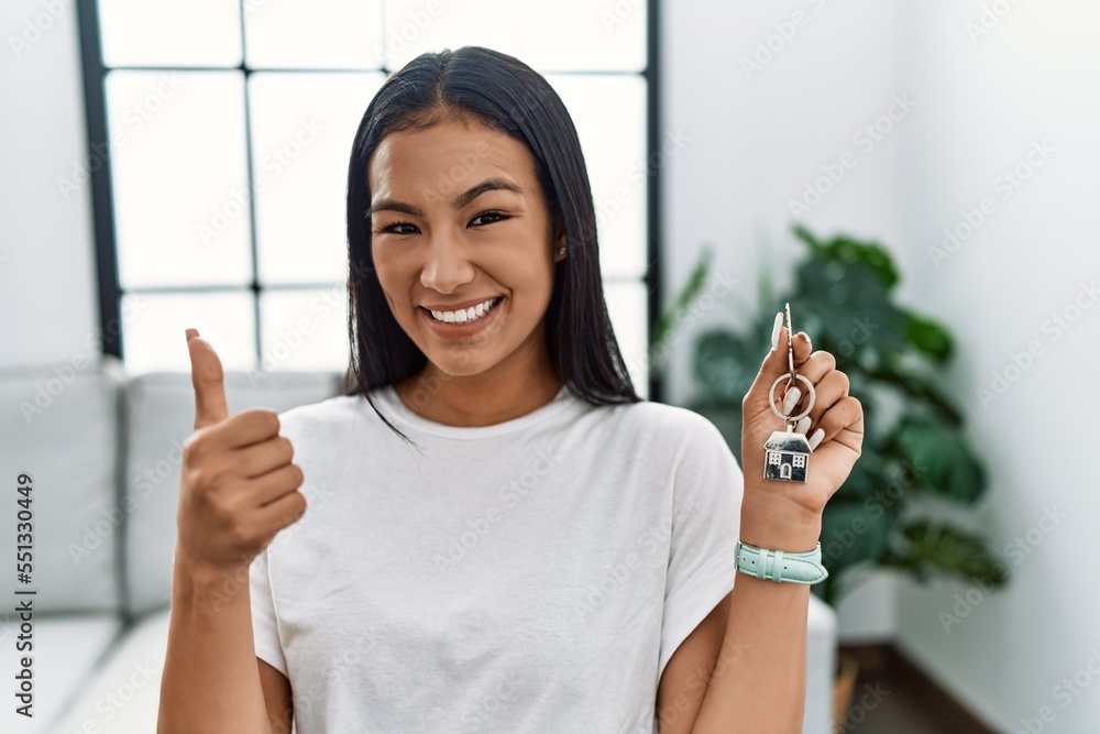 Sticker Young hispanic woman holding keys of new home doing happy thumbs up gesture with hand. approving expression looking at the camera showing success.