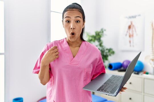 Young hispanic physiotherapist woman using computer laptop at medical clinic scared and amazed with open mouth for surprise, disbelief face