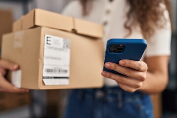Young beautiful hispanic woman ecommerce business worker using smartphone holding package at office