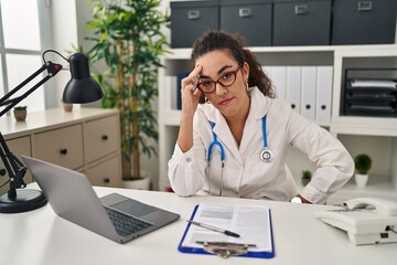 Young hispanic woman wearing doctor uniform and stethoscope worried and stressed about a problem with hand on forehead, nervous and anxious for crisis