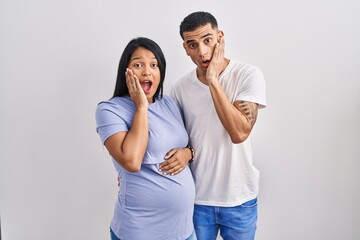 Young hispanic couple expecting a baby standing over background afraid and shocked, surprise and amazed expression with hands on face