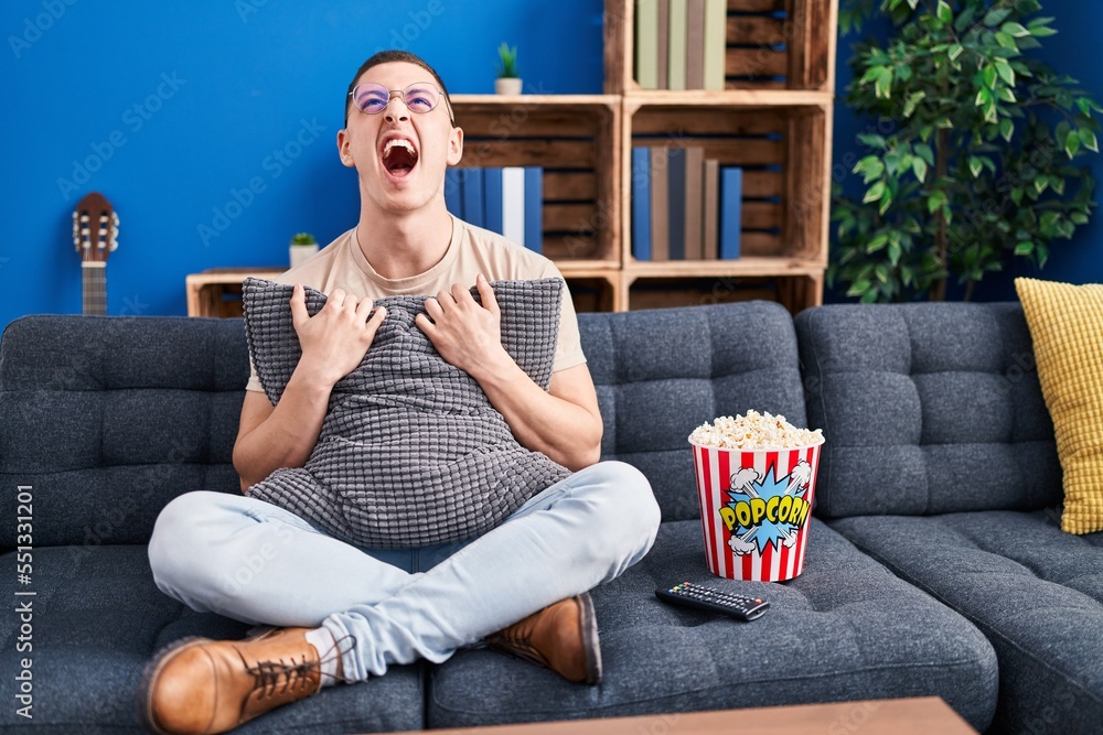 Poster Young man eating popcorn angry and mad screaming frustrated and furious, shouting with anger looking up.