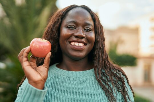 Young african woman smiling happy holding fresh red apple at the city