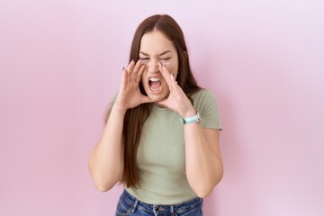 Beautiful brunette woman standing over pink background shouting angry out loud with hands over mouth