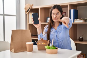 Young brunette woman eating take away food at home showing smartphone screen pointing with finger to the camera and to you, confident gesture looking serious